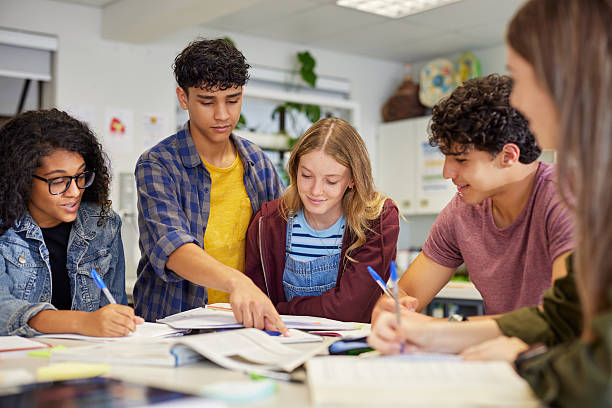 Diverse teenagers students group collaborating during a study session in classroom. Focused high school study group working together with books and notes while preparing for assignment. Girl and guy sharing ideas and helping each other in class at secondary school.